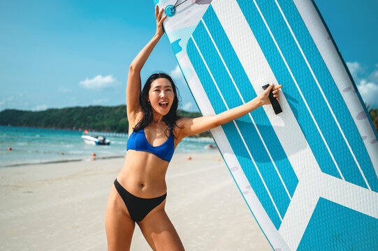 Fit Asian woman posing with a stand-up paddleboard on a sunny tropical beach