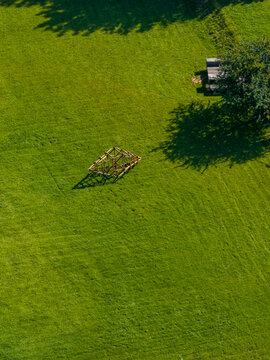 Aerial view of a wooden roof frame structure standing in a lush green field near a tree and a small shed in Raudondvaris, Kaunas County, Lithuania.