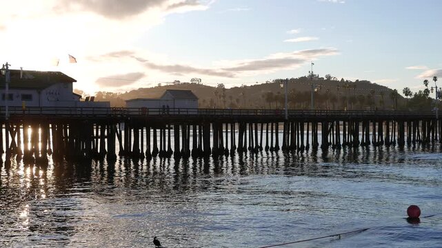 Santa Barbara, California - January 2024: Palm trees, recreationists and tourists walk the pier and enjoy the beautiful weather. Sunset with sun flare.