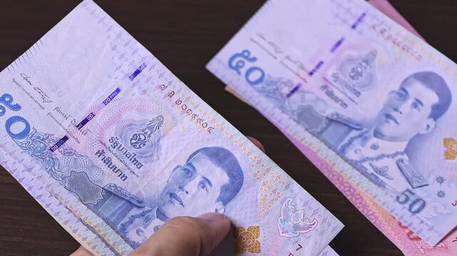 Woman's hands count Thailand baht notes (THB) by placing bills on a dark brown table. Thai cash money of 20, 50, 100, 500 and 1000 baht denominations. Real time motion video.