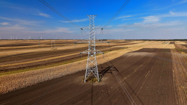 Aerial view of a large metal power transmission tower standing in a vast agricultural field with distant wind turbines under a clear blue sky in Alibunar, Vojvodina, Serbia.