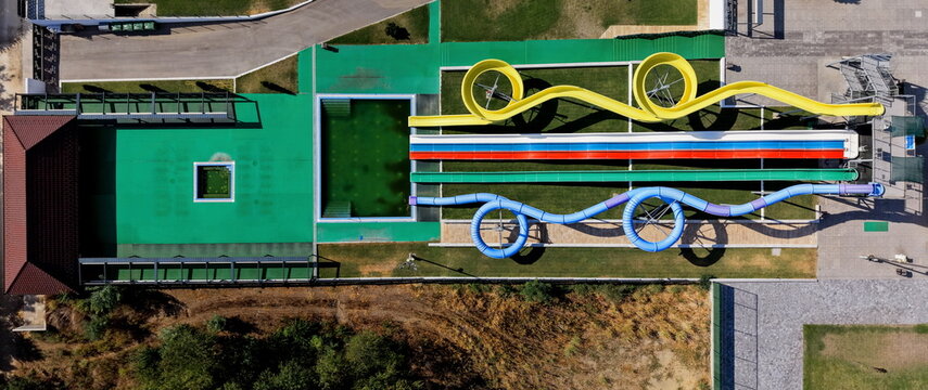 Aerial view of a water park featuring colorful winding slides and a swimming pool surrounded by green decking and trees in Alibunar, Vojvodina, Serbia.
