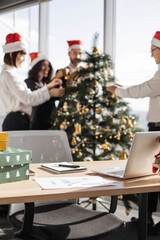 Fototapeta premium Colleagues in Santa hats decorate a Christmas tree in a modern office setting with gifts and a laptop on a desk