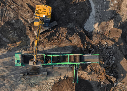 Aerial view of a yellow excavator loading soil into a green mobile screening plant at a construction site in Enschede, Overijssel, Netherlands.