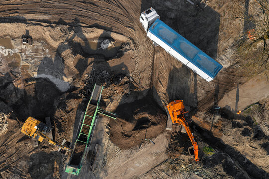 Aerial view of construction machinery including an excavator, a truck, and a soil screening machine working on a dirt site in Enschede, Overijssel, Netherlands.