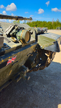 British Warrior infantry fighting vehicle with its main cannon extended, showing tracks and the Union Jack flag detail, parked on a dirt road near a forest under a bright blue sky