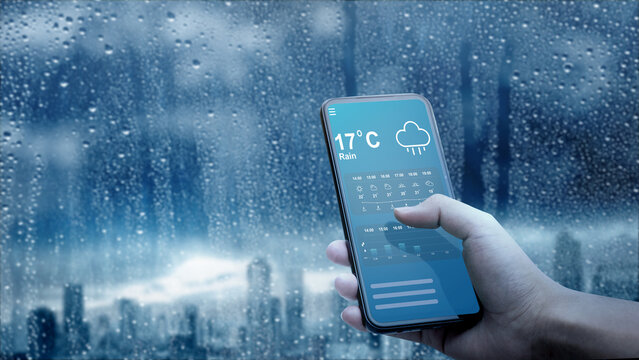Hand holding a smartphone displaying a rain weather forecast app against a wet, rainy window with a blurred city skyline. Concept of Weather