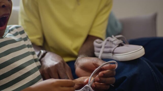 African American father patiently teaching young child how to tie shoelaces at home. Footage suitable for parenting education, fatherhood campaigns and family lifestyle advertising