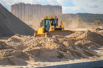 Heavy yellow bulldozer leveling sand mound on a road construction site with dust in the air and high-rise residential building in background. © DmitryRider