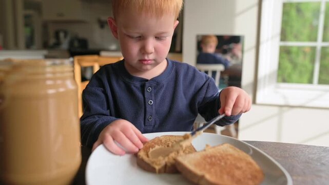 Young boy carefully spreads peanut butter on a slice of toast at a kitchen table, with a jar of peanut butter and sunlight illuminating the scene