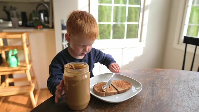 Young boy carefully spreads peanut butter on slices of whole grain bread at a wooden table in a sunlit kitchen with green trees outside