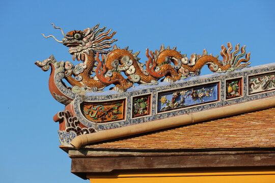 Traditional orange dragon sculpture on crested roof ridge with rectangular flower panels, ancient oriental ornamental rooftop finial architecture.