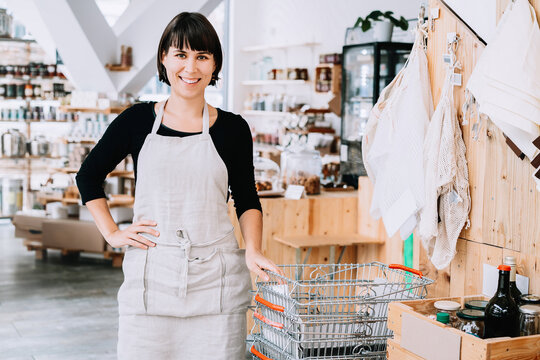 A confident caucasian female entrepreneur stands near metal baskets inside her modern grocery boutique. Environmentally responsible business and eco lifestyle concept.