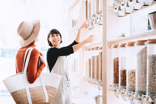 A female shopkeeper in an apron assists a young woman in using gravity food dispensers in a modern shop. Conscious consumer behavior and package free shopping concept.
