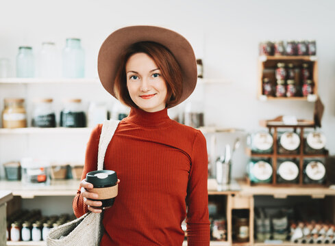 A stylish caucasian female customer wearing a hat holds a reusable coffee cup inside a bulk grocery shop. Mindful lifestyle and conscious environmental consumerism concept.