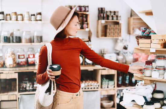 A stylish caucasian female customer wearing a hat holds a reusable coffee cup inside a bulk grocery shop. Mindful lifestyle and conscious environmental consumerism concept.