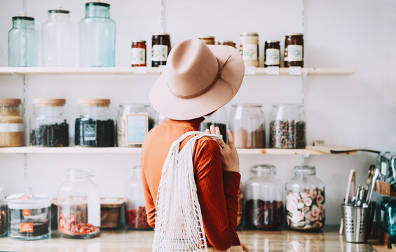 Shopper stands with her back to the camera, looking at organic goods on wooden shelves inside a bright modern store interior. Package free retail environment and conscious consumption concept.