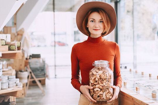 A happy caucasian female customer in a terracotta sweater holds a large glass jar of dried apples inside a bulk shop. Sustainable consumer choices and low waste lifestyle concept.
