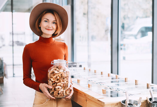 A happy caucasian female customer in a terracotta sweater holds a large glass jar of dried apples inside a bulk shop. Sustainable consumer choices and low waste lifestyle concept.