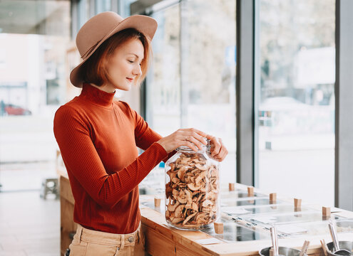 A happy caucasian female customer in a terracotta sweater holds a large glass jar of dried apples inside a bulk shop. Sustainable consumer choices and low waste lifestyle concept.