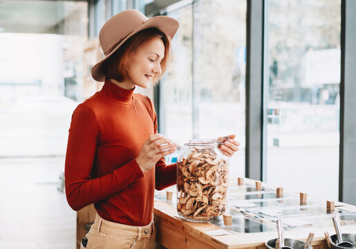 A happy caucasian female customer in a terracotta sweater holds a large glass jar of dried apples inside a bulk shop. Sustainable consumer choices and low waste lifestyle concept.