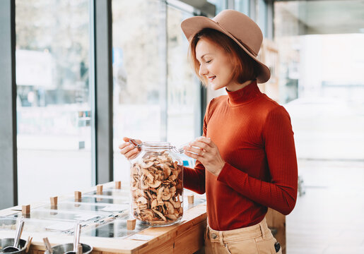 A happy caucasian female customer in a terracotta sweater holds a large glass jar of dried apples inside a bulk shop. Sustainable consumer choices and low waste lifestyle concept.