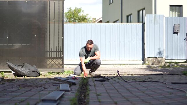 Man crouching to clean and clear a clogged trench drain in the driveway of a residential house with a static camera shot.
