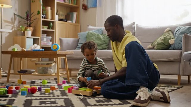 African American father playing with toddler son, assembling wooden toy blocks at home. Clip suitable for parenting campaigns, early childhood education, family lifestyle advertising