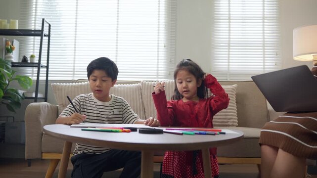 Two asian siblings drawing with colorful markers while their mother works on a laptop nearby. Family life and remote work concept