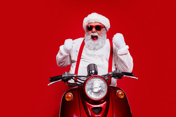 Naklejka premium Excited Santa in red sunglasses and suspenders riding a vintage red scooter for Christmas celebration and holiday fun