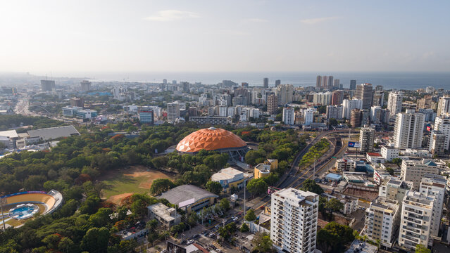 Aerial view of the Palacio de los Deportes Virgilio Travieso Soto with its orange dome, surrounded by trees, urban buildings, and streets in Santo Domingo, Distrito Nacional, Dominican Republic.