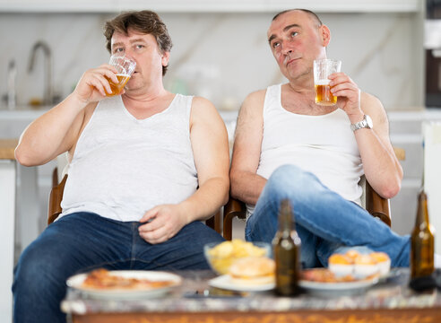 Two adult male friends drinking beer and eating chips at home. Bad habits, eating disorder, alcoholism