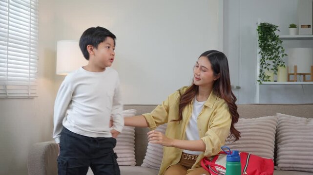 Asian mother helping her son get dressed for school in the living room while preparing his backpack for the day