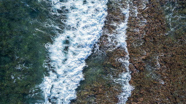 Aerial view of ocean waves crashing against a rocky reef shoreline with clear turquoise water and white sea foam in Bajawa, Nusa Tenggara Timur, Indonesia.
