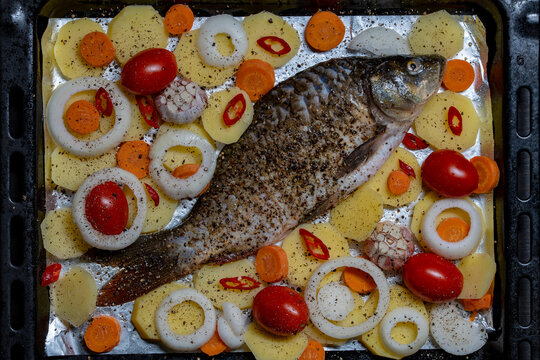Raw whole carp fish with seasoned vegetables on baking tray. Fresh uncooked river fish prepared for baking with potatoes, tomatoes, carrots, onions, garlic and chili pepper on foil, top view