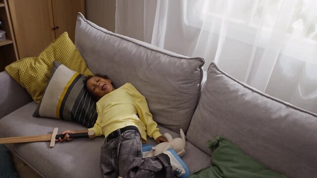 African American child sword fighting with father using toy swords and falling playfully on couch. Perfect for fatherhood campaigns, parenting blogs, and family lifestyle ads