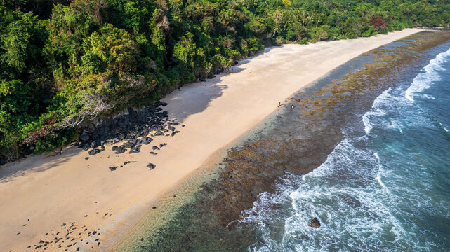 Aerial view of a sandy beach bordered by lush green forest and dark volcanic rocks, with two people walking along the shore in Bajawa, Nusa Tenggara Timur, Indonesia.