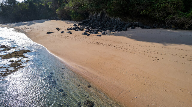Aerial view of a pristine sandy beach with clear turquoise water, dark volcanic rocks along the shoreline, and lush tropical vegetation in Bajawa, Nusa Tenggara Timur, Indonesia.