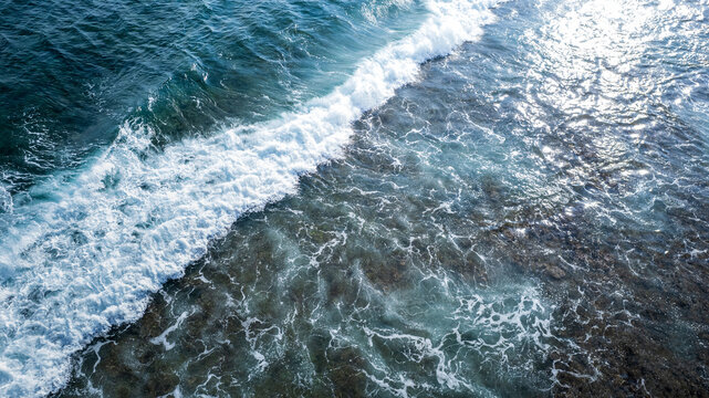 Aerial view of ocean waves breaking over a shallow coral reef with sunlight reflecting on the clear blue water in Bajawa, Nusa Tenggara Timur, Indonesia.