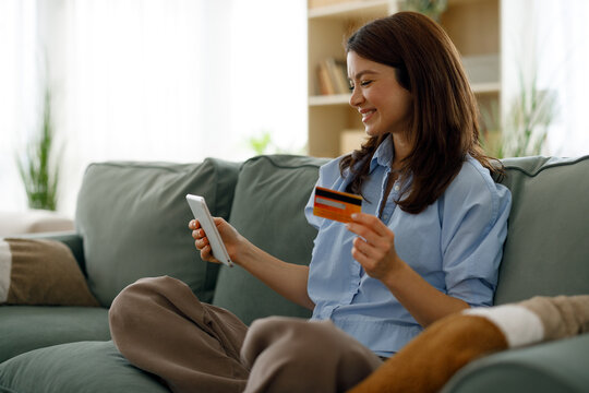 Woman making secure online payment with credit card and mobile phone, shopping on e-commerce application from home couch, smiling while using digital banking service