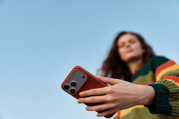 Woman holding smartphone in hand against blue sky. Low angle view with focus on phone. Concept of...