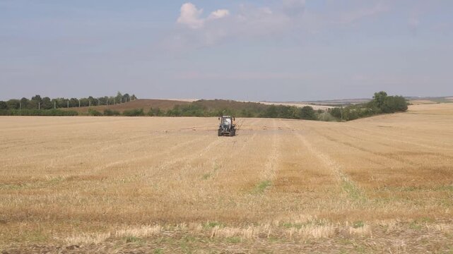 Tractor with a wide crop sprayer working across a golden stubble field in rural Romania, surrounded by rolling farmland and treelines under a soft blue sky.