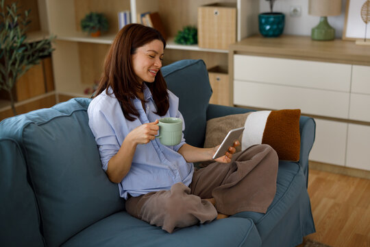 Woman relaxing on a blue sofa, holding a digital tablet and a mug of coffee, enjoying a casual moment of connectivity and comfort in her cozy living room at home