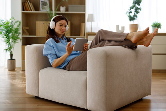 Woman reclining in a cozy armchair at home, barefoot and relaxed, wearing wireless headphones and using a tablet to stream music, browse content, and unwind peacefully