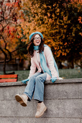 Young woman in pastel coat and beret laughing while sitting on a stone wall in an autumn park with...