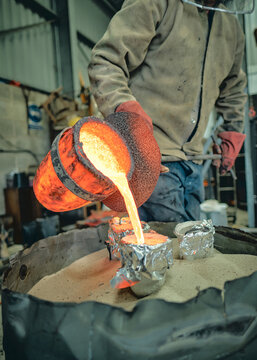 A foundry worker pours intensely glowing molten metal from a heat-scorched crucible into small casting moulds, capturing the raw heat, texture and physical craft 