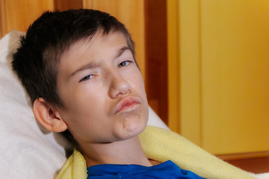 A close-up portrait of a boy; he is reclining on a pillow with a playful, pouting expression; a cozy indoor setting in the background.