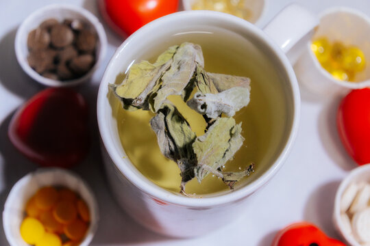 A cup of light peppermint herbal tea with dried leaves on top, surrounded by various tablets, capsules, and red heart shapes, symbolizing natural wellness and complementary healthcare.