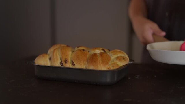 Hand brushing egg wash onto a freshly braided raisin bread loaf in a baking pan, preparing for baking. Homemade sweet bread