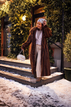 Woman wearing a brown fur coat and patterned pants walks down snowy steps, surrounded by festive greenery and warm lights during winter evening